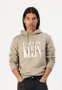 Sudadera beige con un logotipo frontal que dice "CALVIN KLEIN JEANS." Cuenta con capucha con cordón y puños de canalé. Se lleva con jeans oscuros.