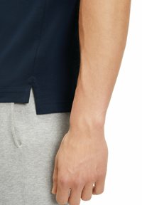 Granatowa koszulka polo z usztywnionej tkaniny, z bocznymi rozporkami, w połączeniu z jasnoszarymi spodniami dresowymi. Ręka oparta na spodniach.