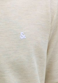 Kreemikas vahvlikootud kangas, millel on väikene valge tikitud "&" sümbol, mis asub rõiva rinnaosa keskel.