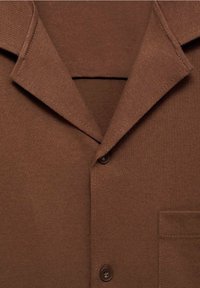 Camiseta de punto marrón con cuello puntiagudo, dos botones en la parte delantera y un bolsillo en el pecho en el lado derecho.