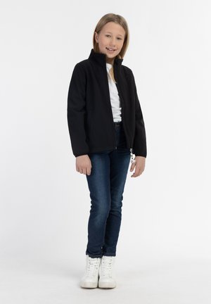 Schwarze Softshelljacke mit Reißverschluss, hohem Kragen und elastischen Bündchen, kombiniert mit blauen Jeans und weißen Sneakern vor einem hellen Hintergrund.
