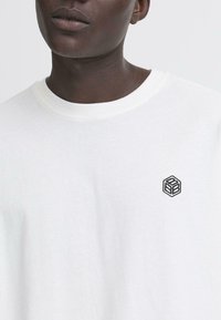 Bílý bavlněný tričko s kulatým výstřihem a krátkými rukávy; obsahuje malý černý vyšitý geometrický logo na levé části hrudi.