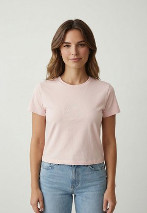 Femme portant un t-shirt rose clair à manches courtes avec des coquillages blancs brodés sur la poitrine, associé à un jean bleu clair, debout contre un mur blanc.