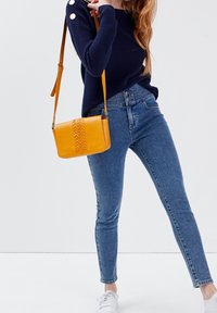 Žlutá kožená kabelka přes rameno s tkaným detailem na přední straně, pravoúhlý tvar, nastavitelný popruh, doplněná modrými úzkými džíny a námořnicky modrým žebrovaným svetrem.