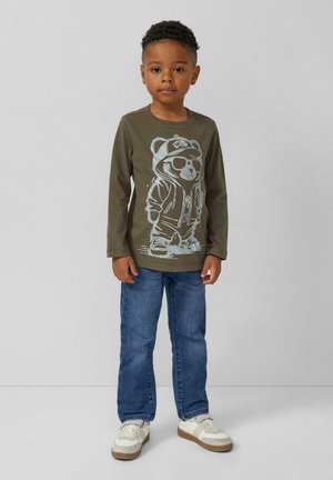 Jongen die staat en een groene lange mouwen shirt met een berenafbeelding draagt, blauwe spijkerbroek en witte sneakers tegen een effen achtergrond.