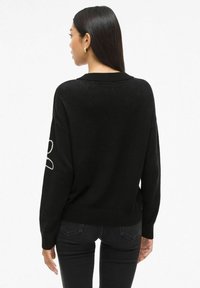 Zwarte trui met een losse pasvorm, lange mouwen en een geribde kraag. Bevat een witte omlijning op de linker mouw. Draagt met donkere jeans.