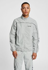 Lysegrå vindjakke med frontlynlås, elastiske manchetter og sort logo. Har kontrasterende sorte sømme og stylingdetaljer på ærmerne.