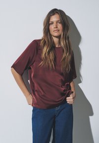 Bordeauxrotes Satin-Kurzarmlshirt mit lockerem Schnitt, kombiniert mit blauen Jeanshosen. Der Stoff hat eine glatte Textur und einen leichten Glanz.