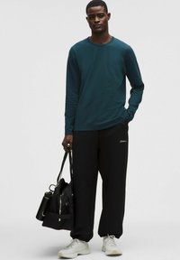 Dlhý rukáv v odtieni teal s voľnými čiernymi nohavicami, zdobený vyšitým logom. Model drží čiernu tašku a má na sebe biele tenisky.