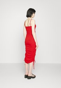 Vestido midi justo vermelho com alças finas e detalhes franzidos nas laterais. Usado com sandálias pretas de salto bloco robusto.