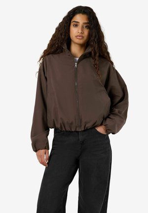 Mujer con cabello largo y rizado que lleva una chaqueta marrón con cremallera y pantalones negros, de pie con una mano en el bolsillo con fondo blanco.