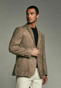Blazer en suédine marron avec un col cranté, fermeture à un bouton et poches avant, associé à une chemise noire et un pantalon clair.