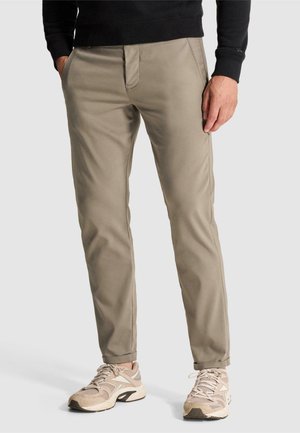 Homme portant un pantalon beige slim, des baskets claires et une chemise noire à manches longues, debout les mains dans les poches.