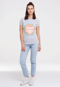 Gray cotton t-shirt with a colorful "Chupa Chups" logo, round neckline, paired with light blue jeans and white sneakers. Simple design.