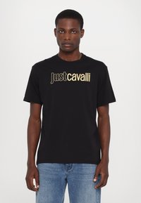 Juodas medvilnės marškinėliai su dideliu auksiniu "Just Cavalli" logotipu prieškrūtinėje dalyje, trumpomis rankovėmis ir laisvu siluetu.