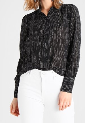 Femme portant un chemisier noir à manches longues avec un motif à pois blancs vertical et un pantalon blanc taille haute, visible du cou jusqu'aux hanches.