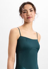 Türkisfarbenes Slipkleid mit dünnen Trägern und quadratischem Ausschnitt, das eine glatte Textur und geometrische Nahtdetails bietet. Transparente Ohrringe setzen einen Akzent.