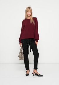 Camisola burgundy de mangas compridas com ombros bufantes, calças pretas com cinto na cintura, sapatos de salto tipo slingback pretos e uma carteira de estampado de pele de cobra.