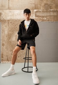 Svart, fluffy zip-up hettegenser over en fargerik graphic t-skjorte, sammen med svarte shorts og hvite joggesko, sittende på en svart barstol.