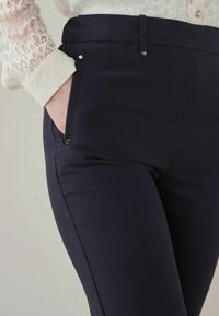 Donna che indossa pantaloni attillati blu scuro con una mano nella tasca laterale e il polsino della camicia in pizzo bianco visibile.