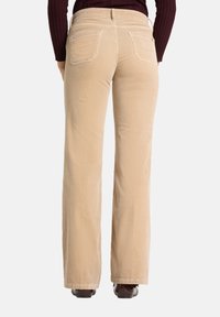 Pantalon en velours côtelé beige à coupe évasée, avec deux poches arrière et une finition texturée, associé à un haut à manches longues foncé.