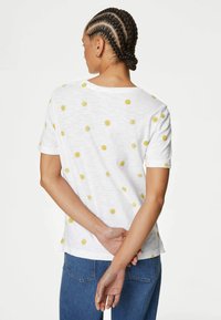 Camiseta blanca de manga corta con patrones de sol amarillos, de corte relajado y escote redondo. Textura suave, combinada con jeans azules.