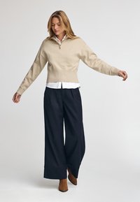 Beżowy sweter dzianinowy z kołnierzem na ćwierć zamka, w zestawie z granatowymi spodniami o szerokiej nogawce i wysokim stanem. Modelka nosi brązowe botki za kostkę.