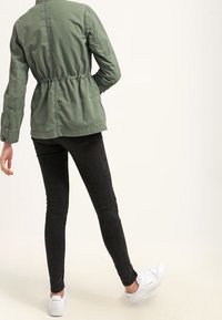Lekka oliwkowa kurtka z elastycznym paskiem w talii, zestawiona z czarnymi obcisłymi dżinsami i białymi trampkami. Tkanina wygląda gładko i swobodnie.