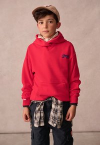 Červená mikina s kapucí a modrým logem, tmavé cargo kalhoty a károvaná flanelová košile uvázaná v pase, doplněné béžovou kšiltovkou.