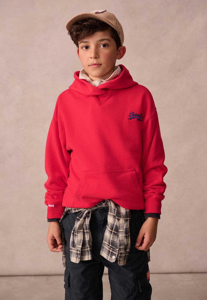 Červená mikina s kapucí a modrým logem, tmavé cargo kalhoty a károvaná flanelová košile uvázaná v pase, doplněné béžovou kšiltovkou.