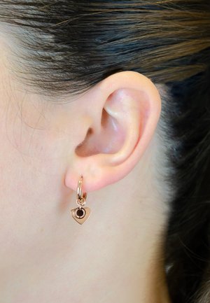 Roségoldene herzförmige Ohrringe mit einem kleinen schwarzen Edelstein, ausgestattet mit einem klappbaren Hoop-Design, neben dem Ohr eines Kunden präsentiert.