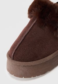 Primo piano di una pantofola in suede marrone per l'inverno, con suola in gomma spessa e bordo in pelliccia sintetica marrone scuro attorno all'apertura.