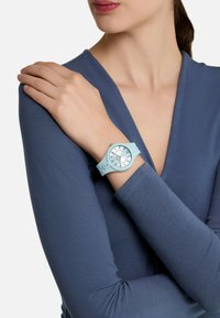 Reloj de silicona azul claro con esfera redonda y acentos plateados, llevado en una mano con una camiseta de manga larga azul marino ajustada. Diseño simple.
