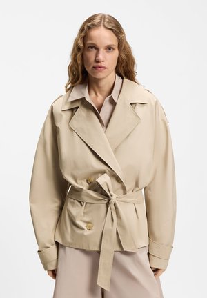 Vaaleat laineikkaat hiukset omaava nainen, jolla on beige vyötäröllä solmittu trenssitakki vaalean paidan päällä, seisoo yksinkertaisen valkoisen taustan edessä.