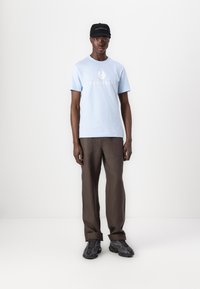 Ljusblå bomull t-shirt med "BELSTAFF"-logo, bruna byxor och svart keps. Outfit kompletteras med svarta texturerade sneakers.