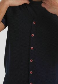 Uomo che indossa una camicia scura a trama con bottoni rossi, mentre aggiusta il colletto con entrambe le mani.