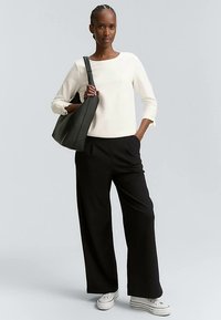 Černé široké kalhoty, bílý texturovaný svetr a bílé tenisky. Modelka drží černou kabelku, stojí před jednobarevným šedým pozadím.