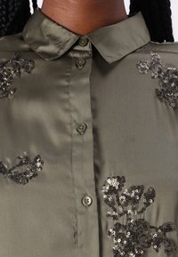 Olīvu zaļš satīna blūze ar izvirzītu apkakli, priekšējiem pogām un dekoratīvu melnu sequīnu ziedu izšuvumu uz krūšu daļas.