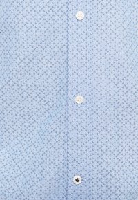 Camicia azzurro chiaro con un sottile motivo a diamante, caratterizzata da piccoli punti blu scuro e bottoni bianchi lungo la parte anteriore. Tessuto morbido.