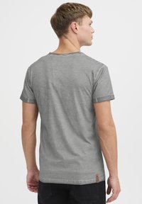 Camiseta gris de manga corta hecha de algodón. Cuenta con un escote redondo, puños enrollados y una pequeña etiqueta de marca en el dobladillo. Textura suave.
