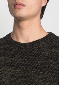 Pull en maille vert foncé avec un col rond et un motif texturé. A un ajustement près du cou et des épaules.