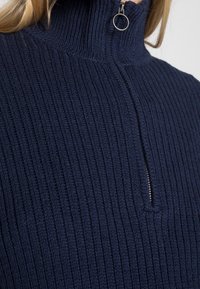 Pull marin côtelé avec col montant et détail de fermeture éclair argentée. Le tissu en maille texturée ajoute de la dimension et de la chaleur.