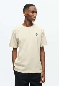 Beige bomulds t-shirt med korte ærmer, rund halsudskæring og lille grøn logo på brystet; afslappet pasform og glat tekstur.