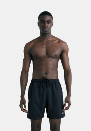 Pantaloni da bagno neri con coulisse in vita, dotati di tasche laterali e un piccolo logo bianco sulla gamba sinistra. Tessuto liscio e leggero.