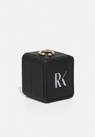 Black leather ring box with a gold ring featuring two small diamond-like accents on top. Box has a white logo "RK" on the front.