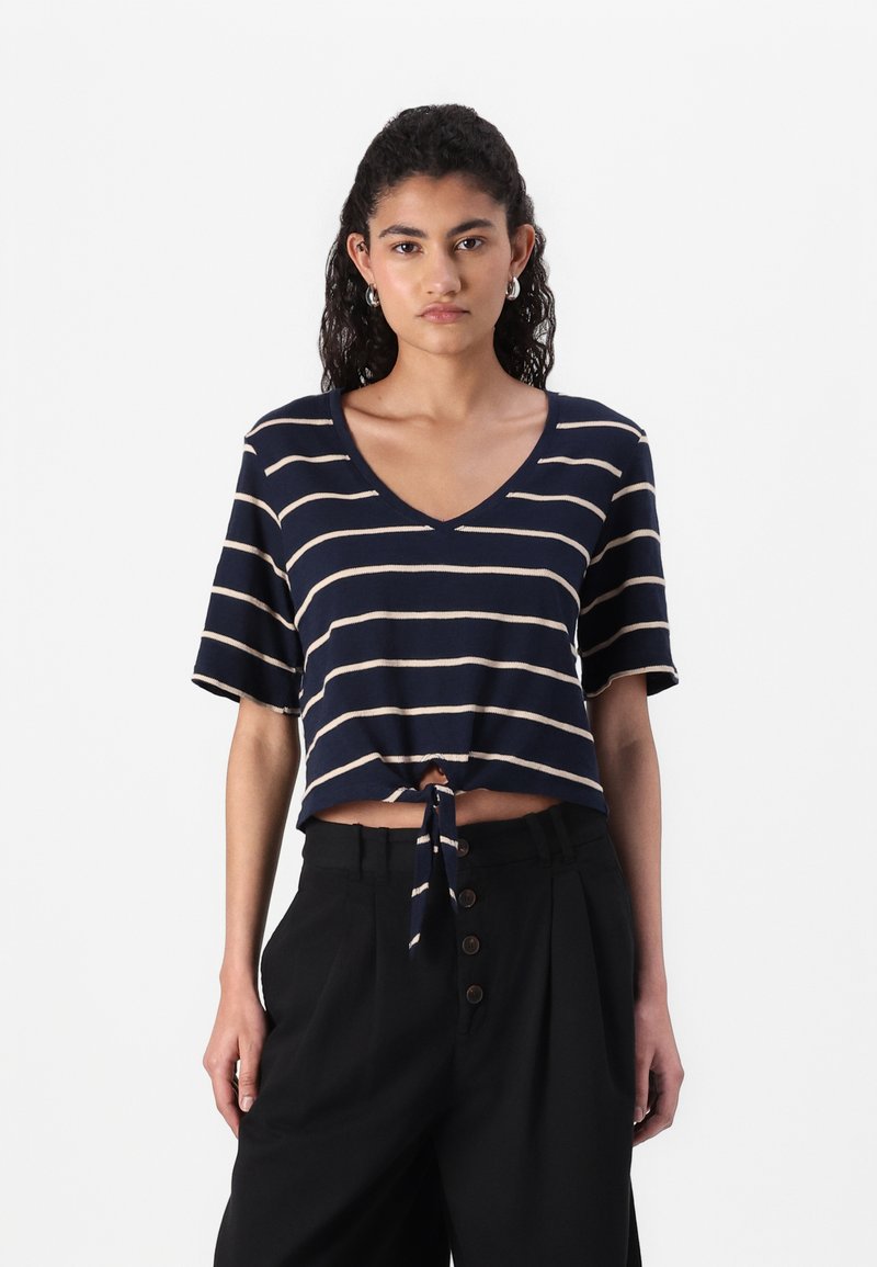 Top corto de color navy con rayas horizontales beige, escote en V, mangas cortas y detalle de lazo en el frente. Combinado con pantalones negros de talle alto con botones.