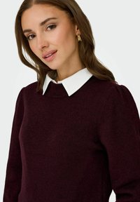 Maglione bordeaux con colletto bianco e girocollo, caratterizzato da una morbida trama a maglia e dettagli sottili sulle spalle. Orecchini dorati completano il look.