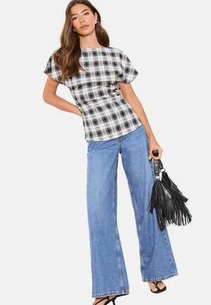 Naine kannab must-valget ruudulist tippu, laiade jalgadega siniseid pükse, musti kontsaga sandaale, hoides musta tutiga käekotti, poseerib valge tausta ees.