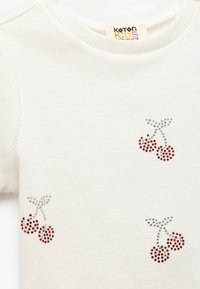Camisa blanca de manga corta con textura acanalada, que presenta un diseño de cereza con rhinestones en rojo y plata en la parte frontal. Etiqueta de Koton Kids visible.