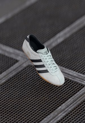 Biały sneaker Adidas z czarnymi paskami i gumową podeszwą unosi się nad metalową kratką, pokazany pod kątem z widocznymi sznurowadłami i szczegółami logo.
